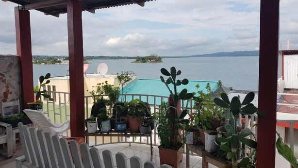 A covered balcony with potted plants, a white chair, and a view of a body of water and distant land. - Hotel Posada Tayazal (Flores)