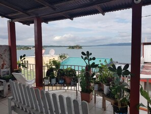 A covered balcony with potted plants, a white chair, and a view of a body of water and distant land. - Hotel Posada Tayazal (Flores)