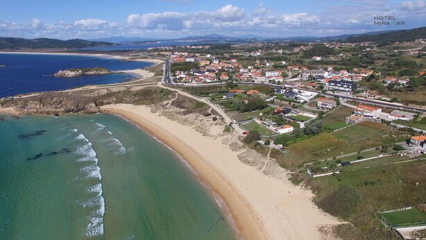 Vista aérea de un pueblo costero con playa, zonas residenciales y una carretera que lleva al mar.