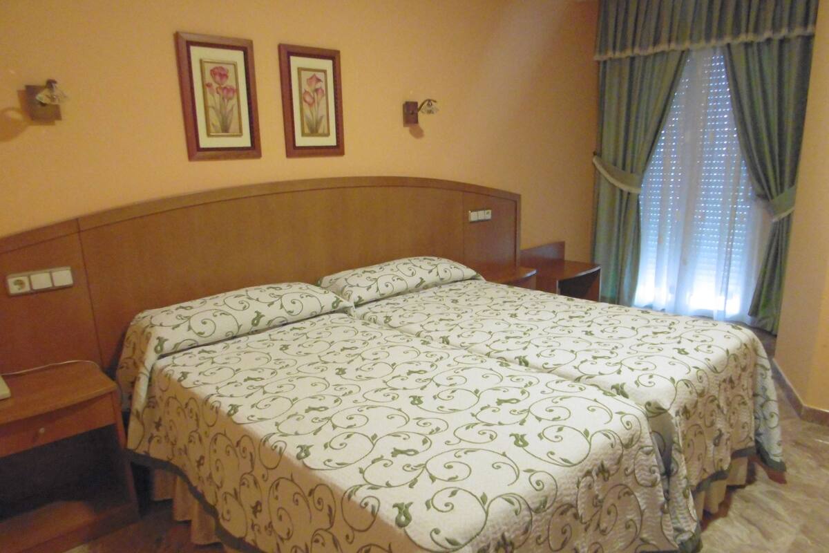 A hotel room with a large bed, two bedside tables, a nightstand, a wooden headboard, and a window with curtains.