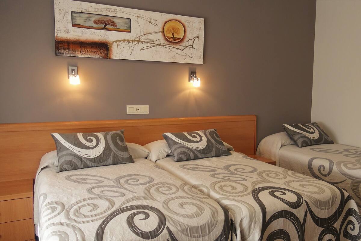 A hotel room with two beds, each with a headboard and a patterned bedspread. There are two wall-mounted lights above the beds and a painting on the wall.