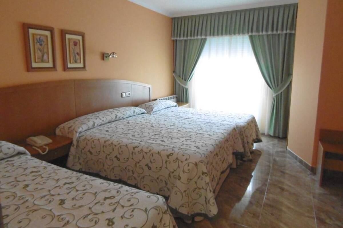A hotel room with three beds, a wooden headboard, a bedside table with a telephone, and a window with curtains.
