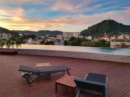 A rooftop terrace with wooden flooring, two lounge chairs, and a pool.