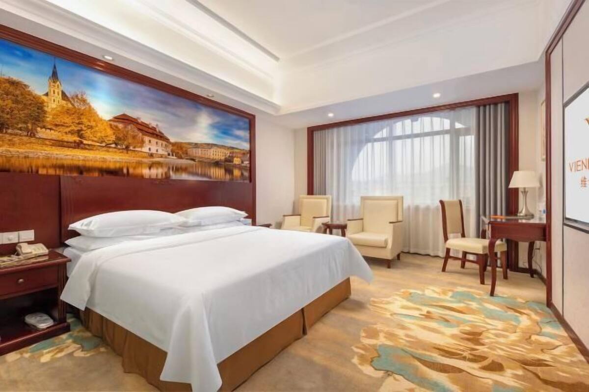 Une chambre d’hôtel avec un grand lit, un bureau, deux chaises, un tableau au mur et une vue sur un immeuble par la fenêtre.