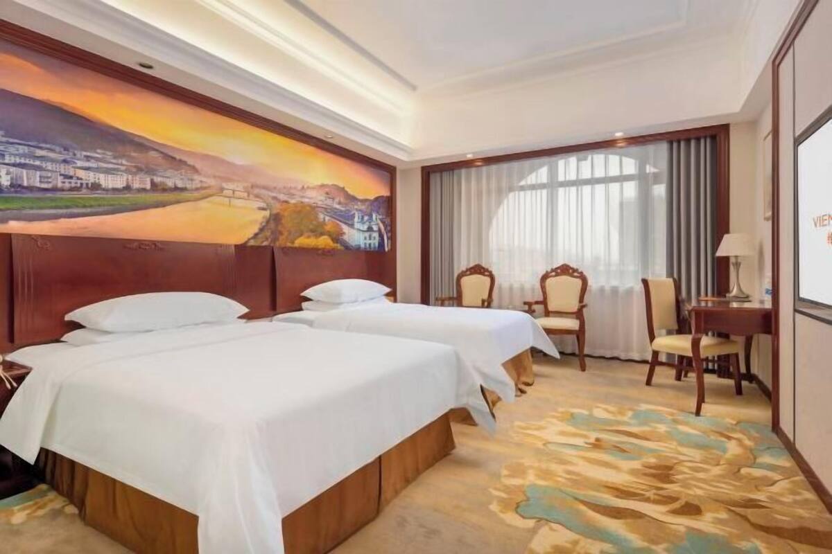 Une chambre d’hôtel avec deux lits, un bureau, des chaises, une grande fenêtre avec des rideaux et un tableau au mur.