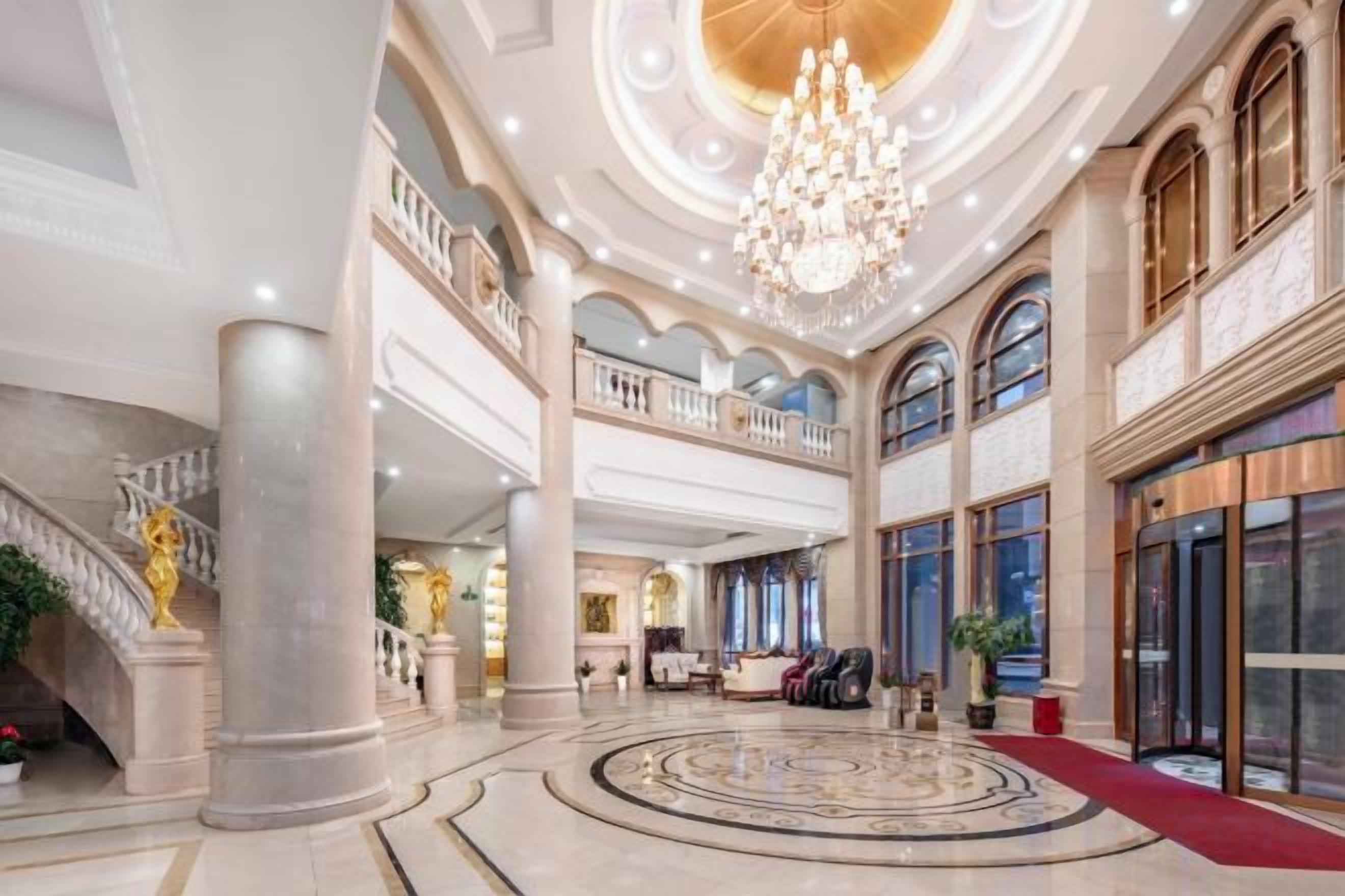 A grand lobby with a central circular floor design, large chandeliers, and a staircase with ornate details.