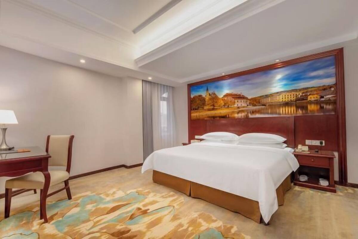 Une chambre d’hôtel avec un grand lit, un bureau avec une chaise, une table de chevet et un tableau représentant un paysage accroché au mur.