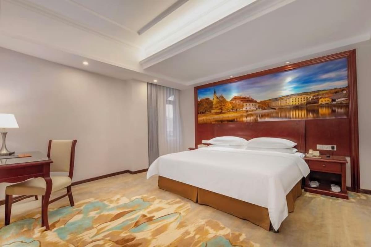 A hotel room with a bed, a desk, a chair, a nightstand, and a painting on the wall.