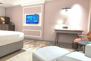 Quarto de hotel com cama, escrivaninha, cadeira, TV fixada na parede e tapete estampado.