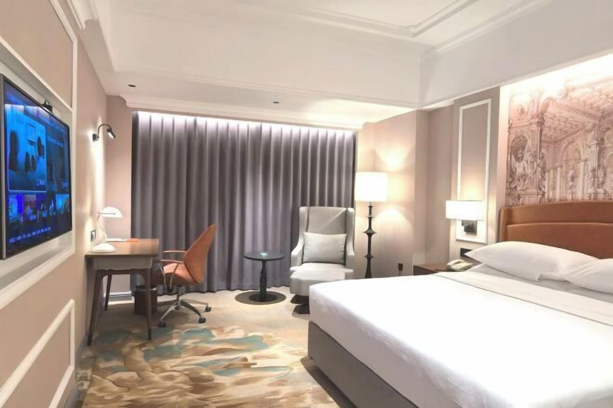 Quarto de hotel com uma cama grande, uma escrivaninha com cadeira, televisão e uma janela com cortinas.