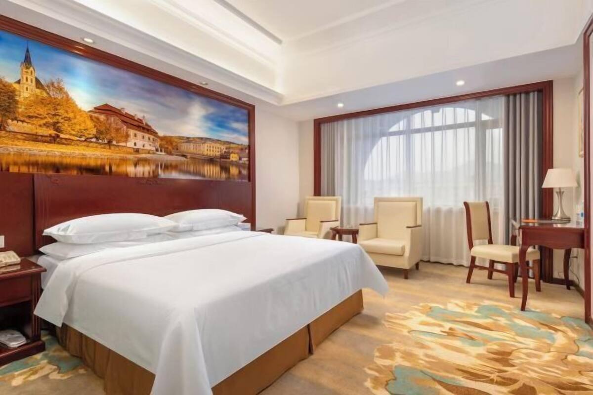 Une chambre d’hôtel avec un grand lit, un bureau avec une lampe, deux fauteuils et un tableau représentant un château accroché au mur.