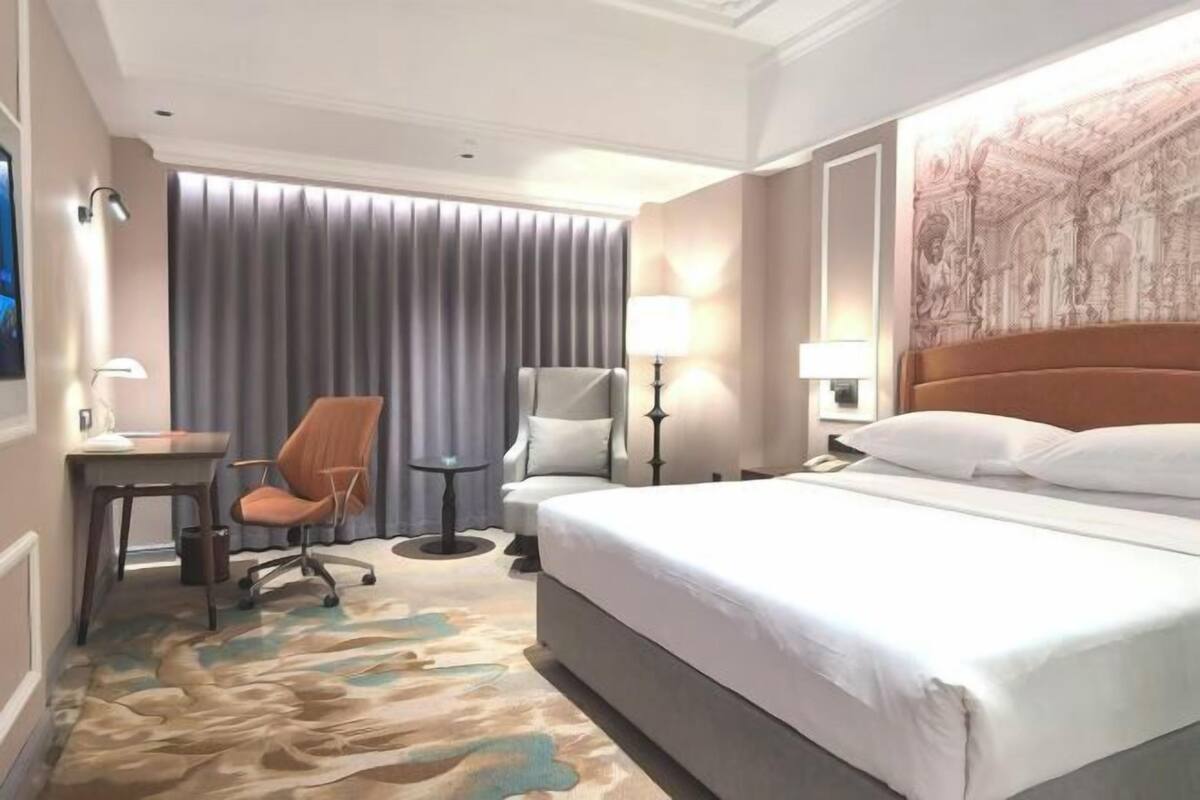 Une chambre d’hôtel avec un lit, un bureau, une chaise, une lampe et une grande tapisserie au mur.