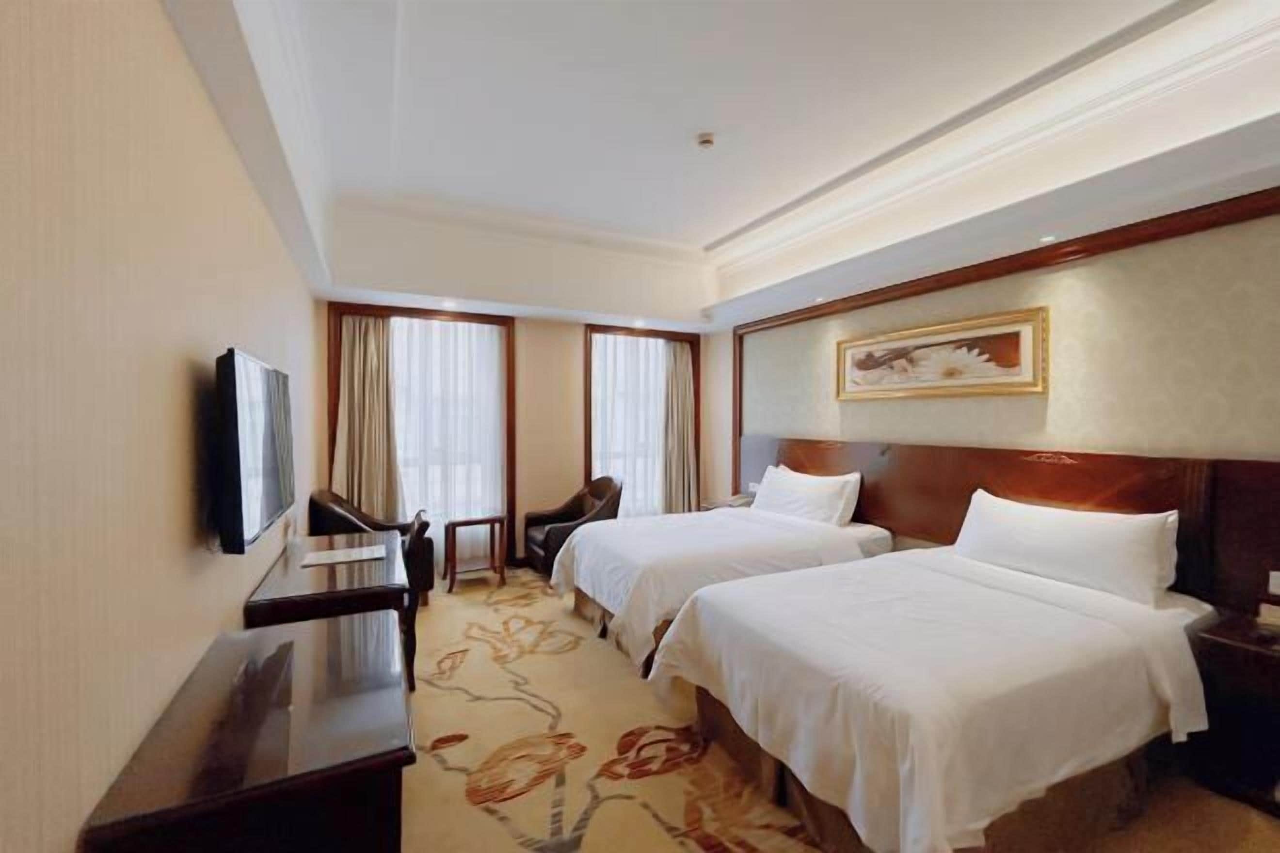 A hotel room with two beds, a desk, a chair, and a television.