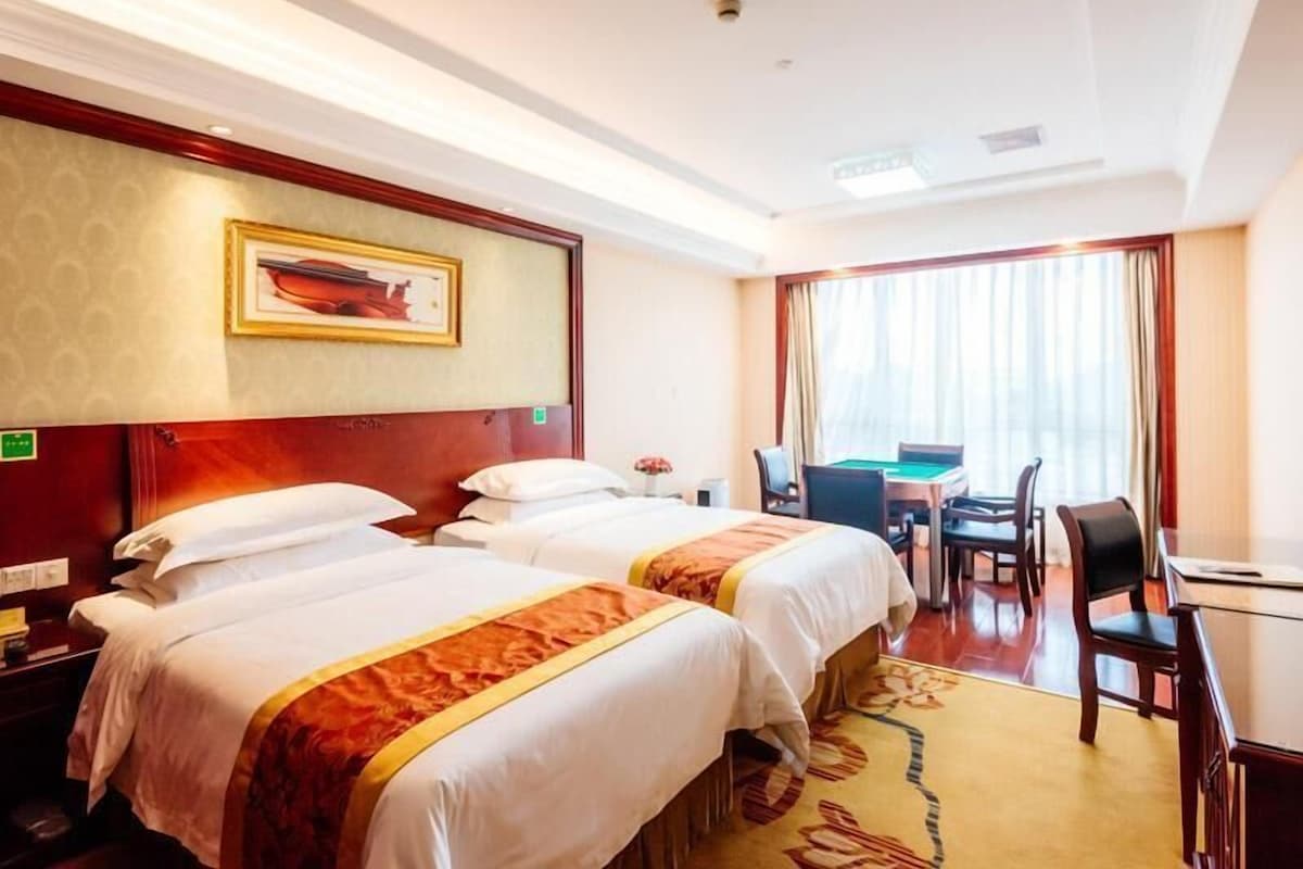 A hotel room with two beds, a desk, a chair, a pool table, and a large window with curtains.
