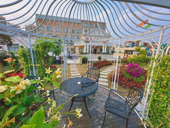 A covered outdoor seating area with a table and chairs, surrounded by plants and flowers.