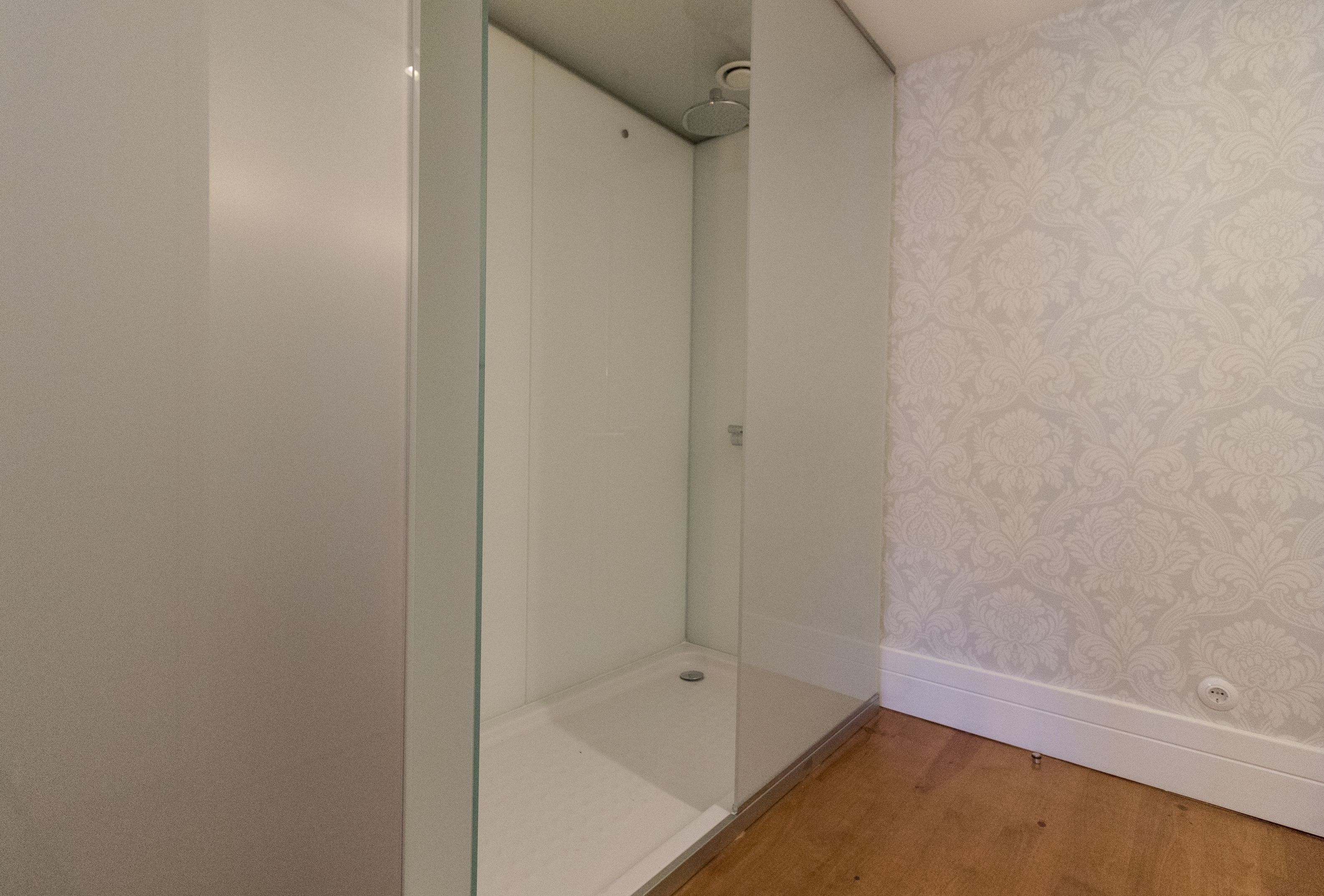Um banheiro com box de vidro, piso de madeira e papel de parede estampado na parede ao lado.