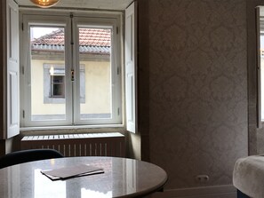 A room with a round table, a window with a view of a building with a tiled roof, and patterned wallpaper. - Casa da Picaria (Porto)
