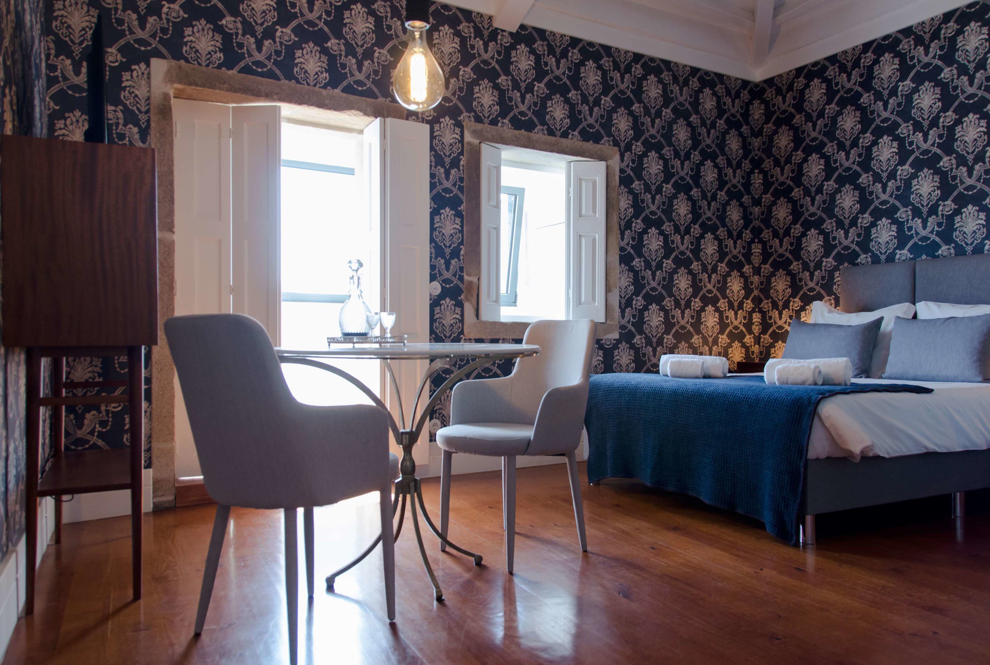 Habitación con cama, escritorio y dos sillas. Se ve una ventana con contraventanas abiertas y una lámpara colgante.