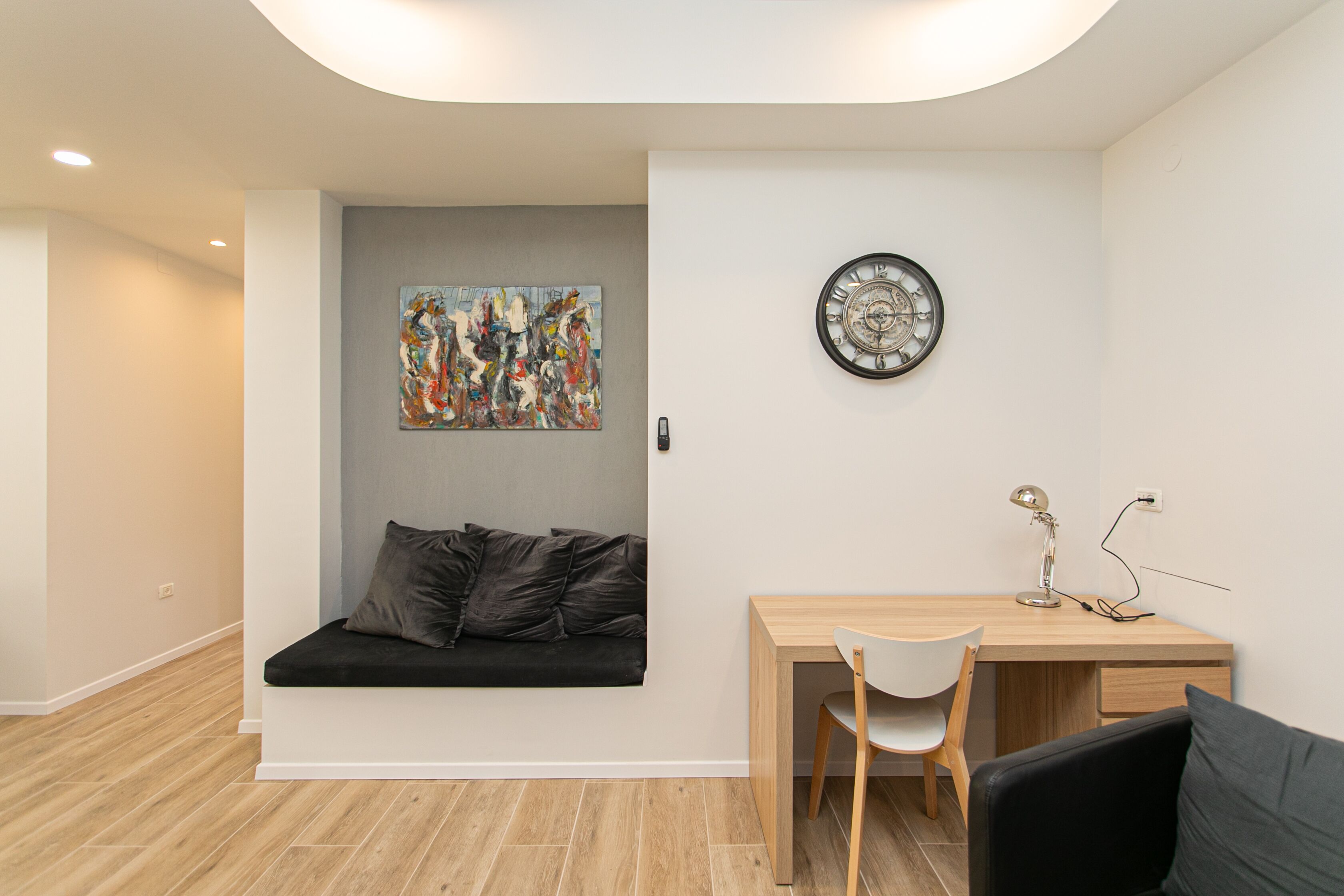 A modern room with a wooden desk, a white chair, a black couch, and a wall clock.