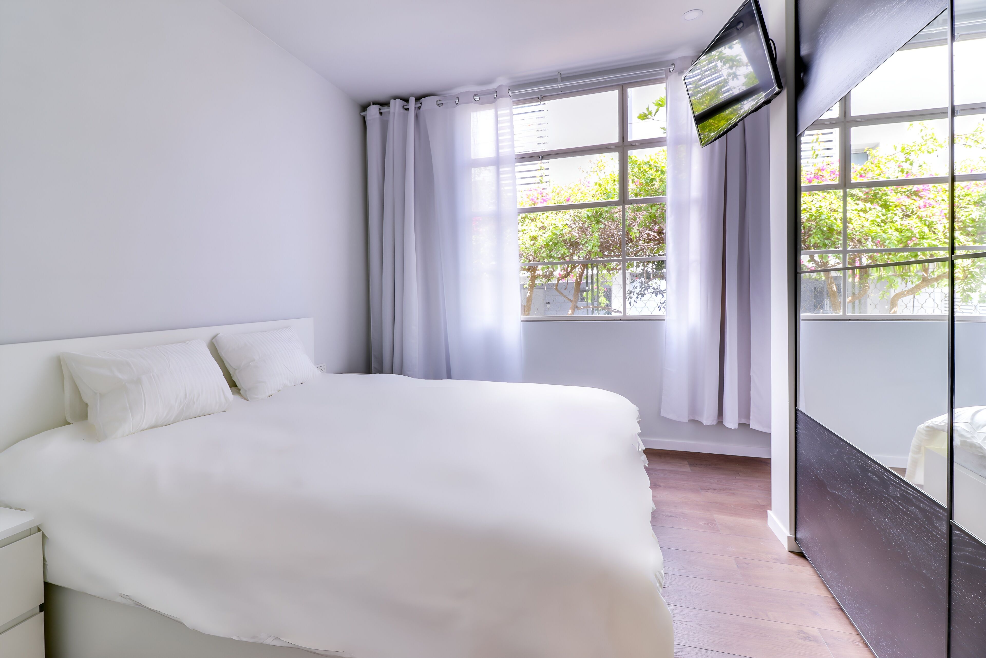 A bedroom with a large bed, a TV mounted on the wall, and a window with curtains.