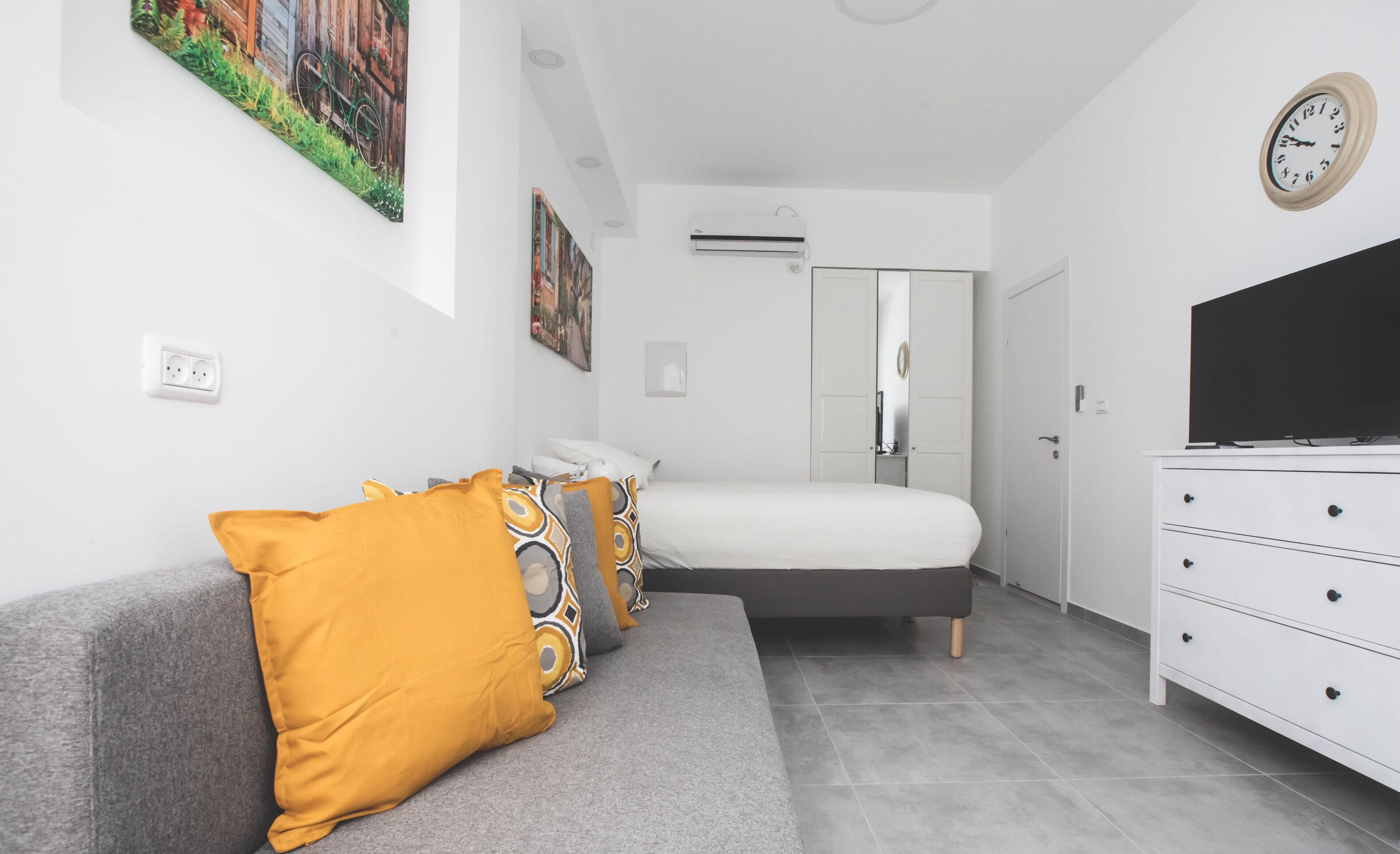 A modern bedroom with a grey sofa, a bed, a dresser, and a television.