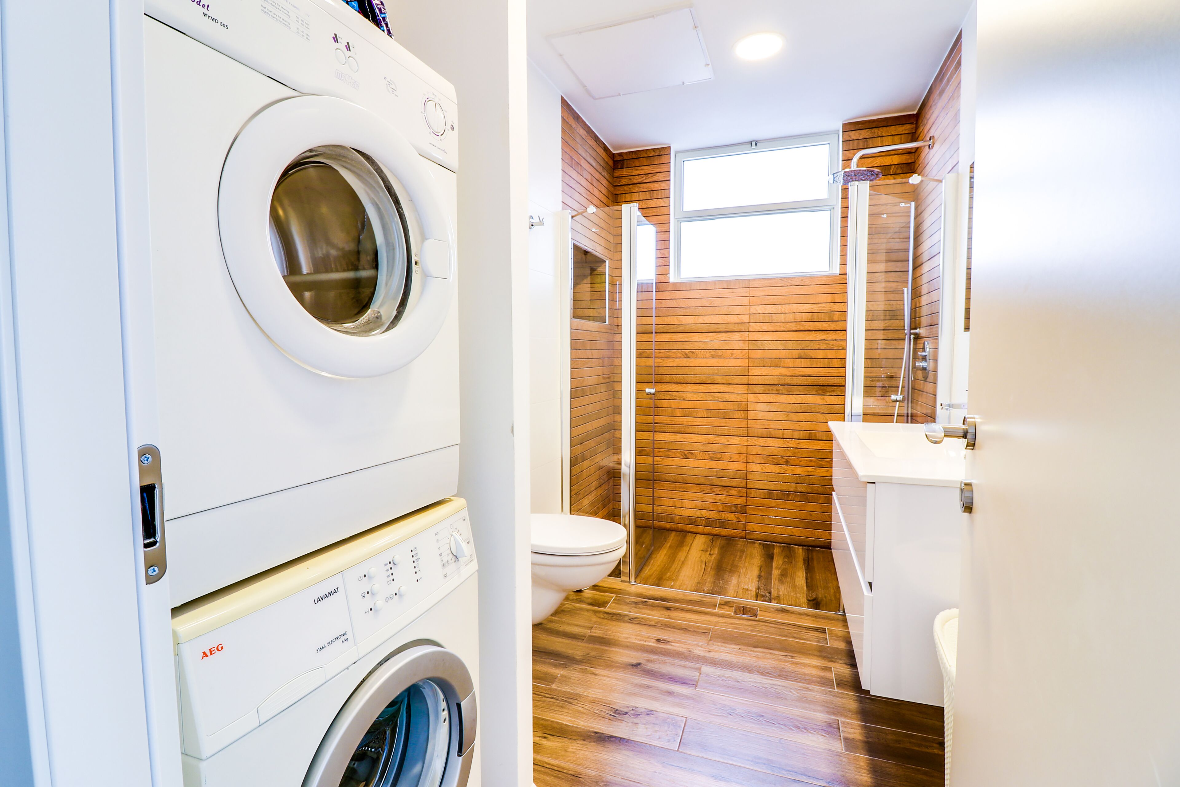 A compact laundry room with a front-loading washing machine and dryer, a wooden-finished shower, and a white toilet.