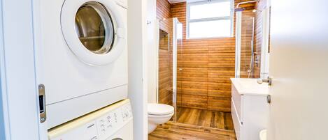 A compact laundry room with a front-loading washing machine and dryer, a wooden-finished shower, and a white toilet.