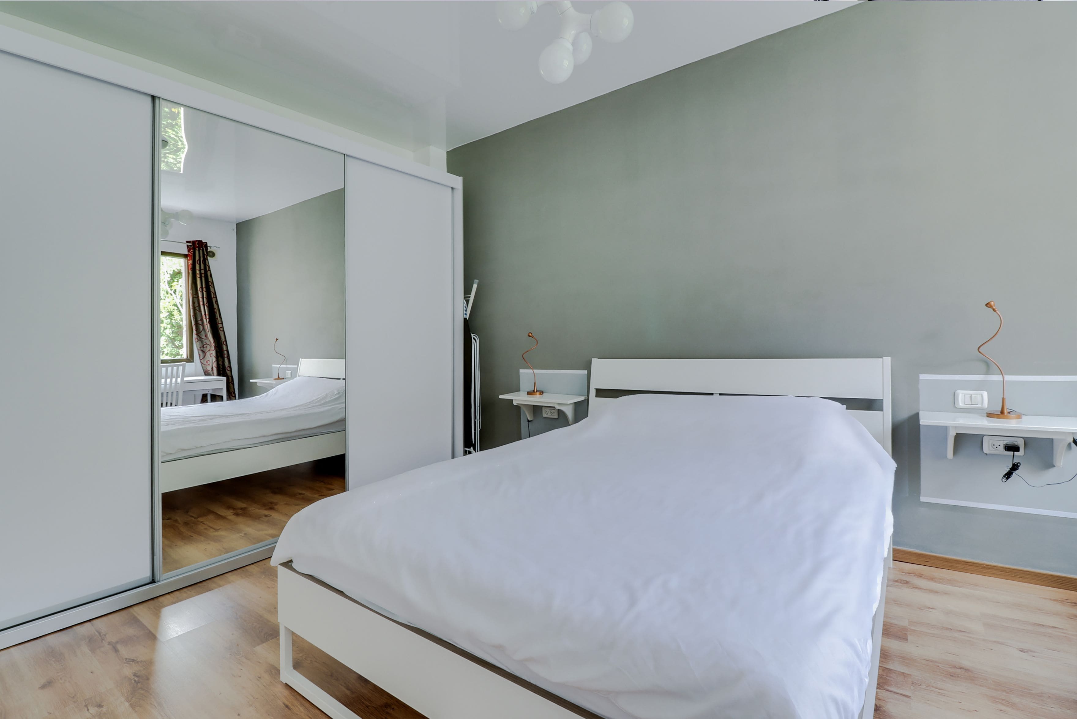 A modern bedroom with a large bed, a wardrobe with a mirror, bedside tables, and a small desk.