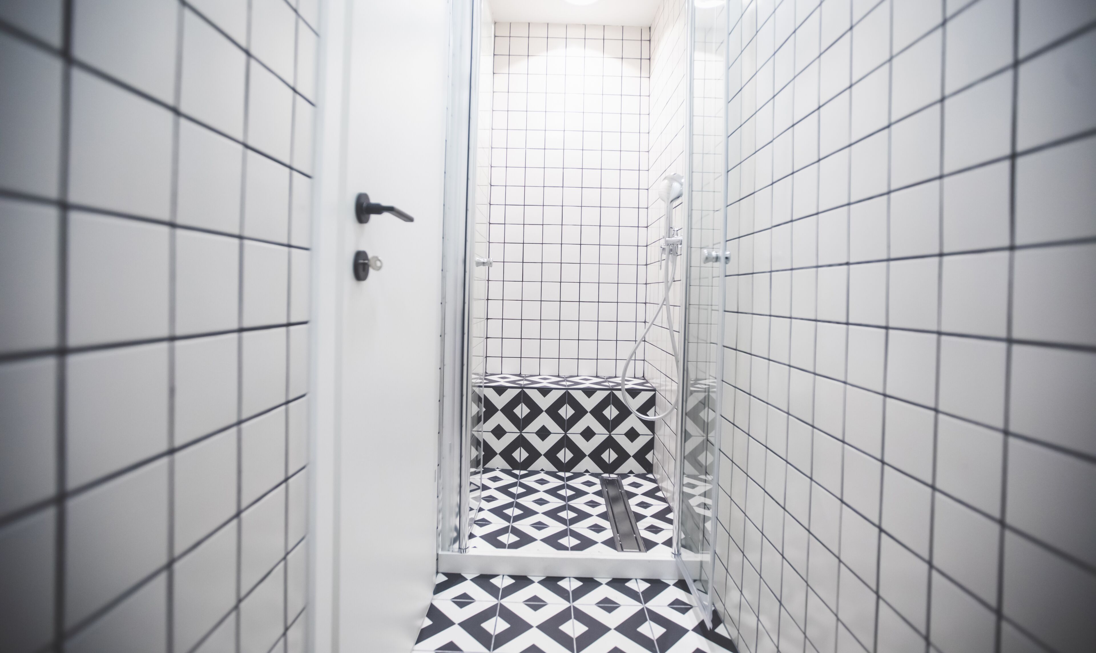 A narrow bathroom with white tiled walls and floor, featuring a geometric patterned tile design on the lower section.