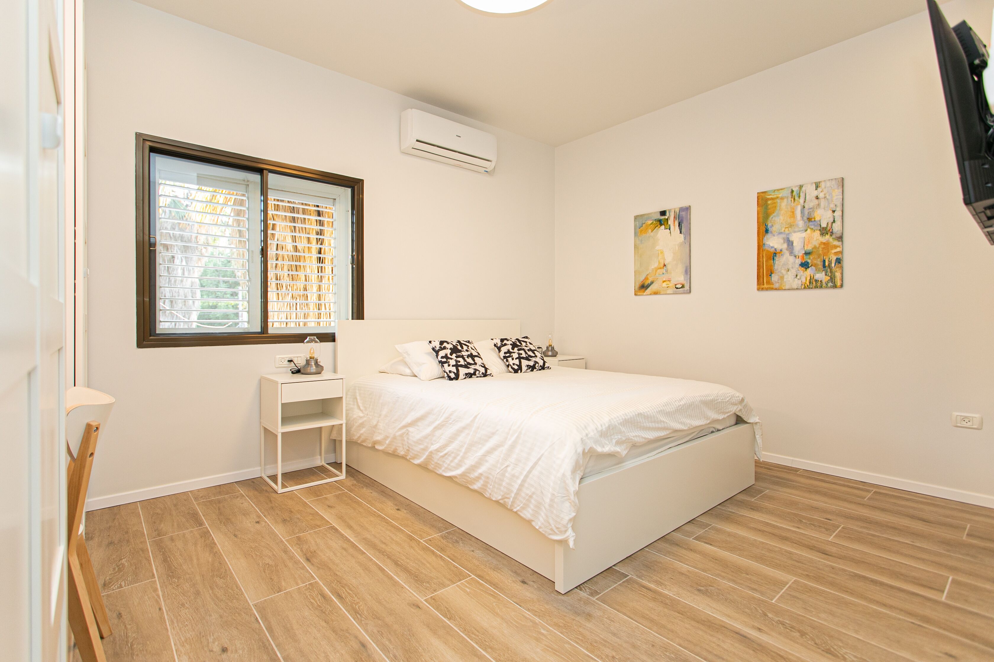 Un dormitorio con cama, mesita de noche, ventana con persianas y dos cuadros en la pared.