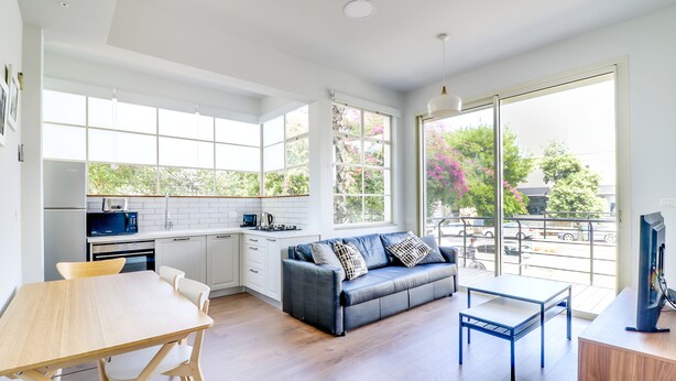 A modern kitchen with white cabinets, a dining table, and a sofa. There is a large window providing a view of the outdoors. - Carmel Boutique (Haifa)