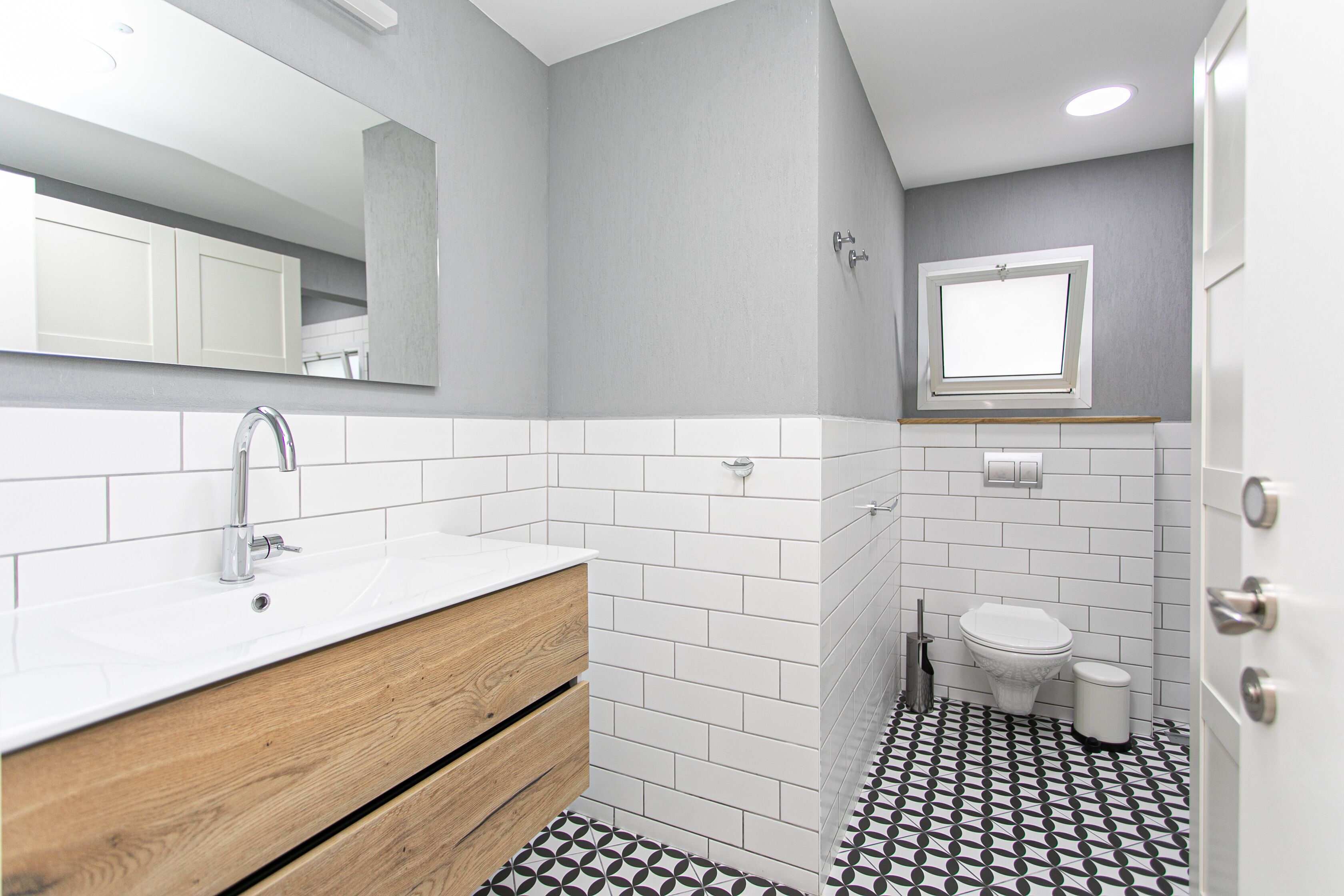 A bathroom with a white sink, wooden vanity, white tiled walls, a window, and a toilet.