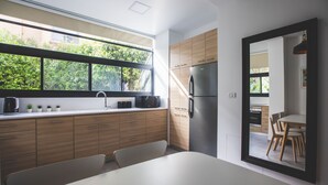 A modern kitchen with wooden cabinets, a stainless steel refrigerator, a microwave, and a sink. There is a window providing a view of greenery outside. - Carmel Boutique (Haifa)