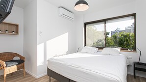 A modern bedroom with a large bed, a wicker chair, a window with a view of greenery, and an air conditioning unit. - Carmel Boutique (Haifa)