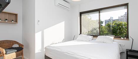 Une chambre moderne avec un grand lit, un fauteuil en osier, une fenêtre donnant sur un paysage verdoyant et un appareil de climatisation.