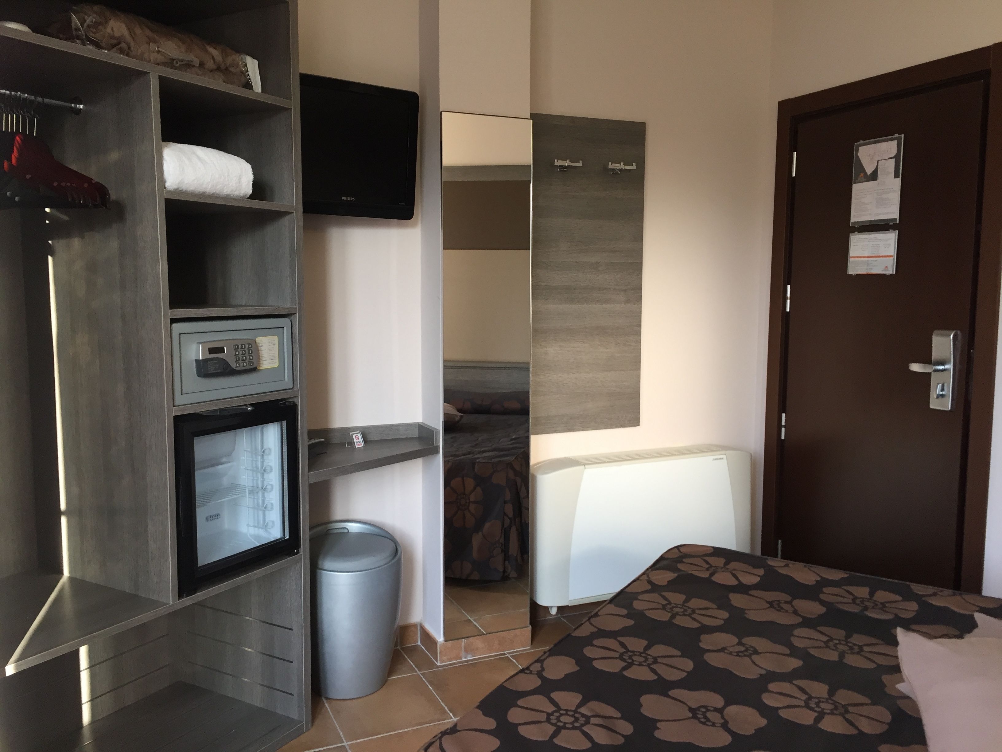 A hotel room with a bed, a wardrobe, a small desk with a computer, a trash bin, and a door with a notice.