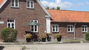 A brick building with a red-tiled roof, multiple windows, and outdoor seating with potted plants. - Hostel Rudbøl (Højer)