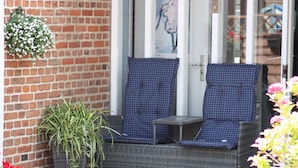 A wicker chair with a checkered cushion, a potted plant, and a hanging flower basket against a brick wall. - Hostel Rudbøl (Højer)