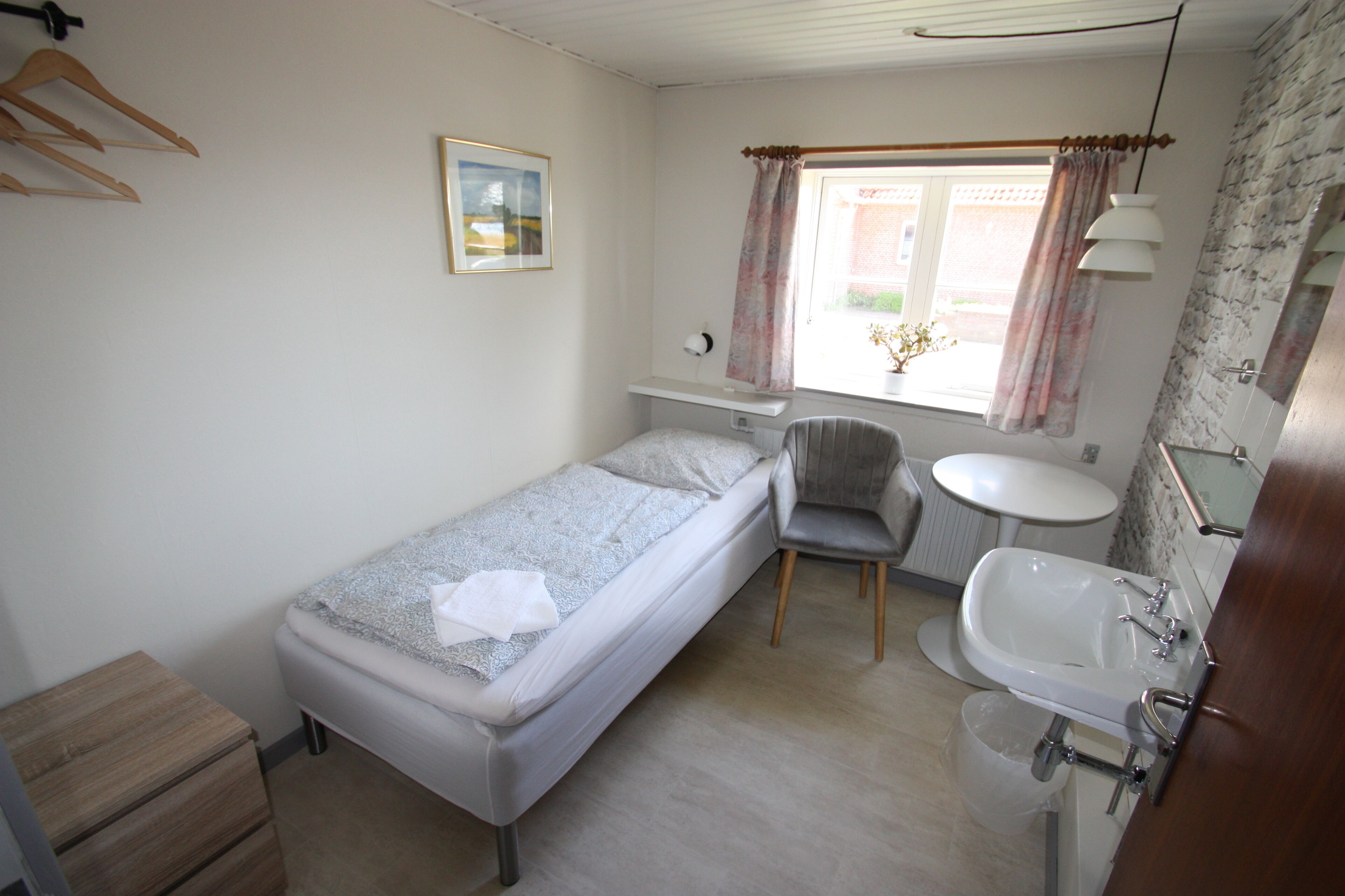 A small, single-bed room with a bedside table, a chair, a round table, and a sink.