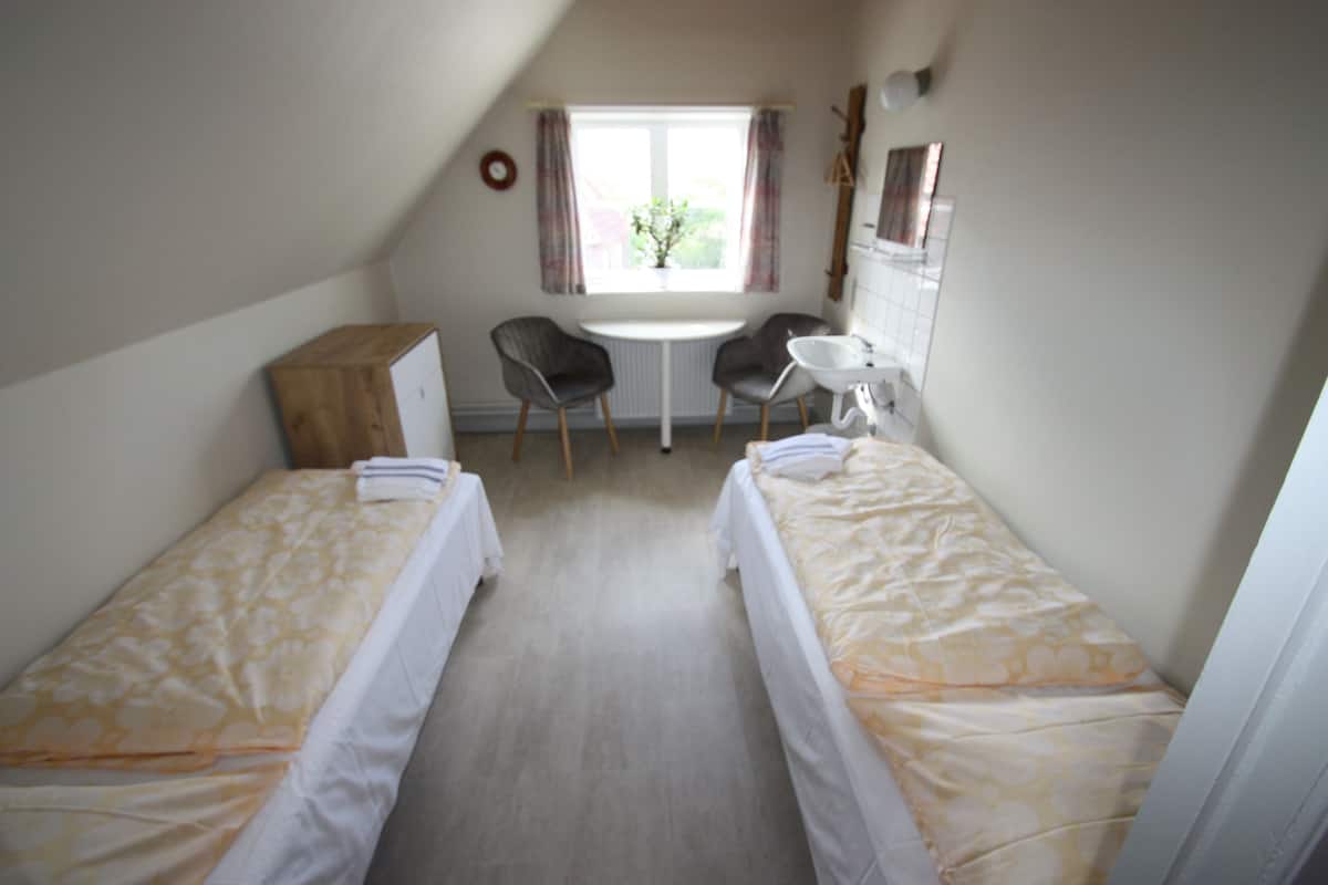 Quarto com duas camas de solteiro, uma mesa pequena, uma cadeira e janela com cortinas.