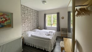 A bedroom with a double bed, a wooden chair, a radiator, a window with curtains, a clock, and a wall with a floral wallpaper. - Hostel Rudbøl (Højer)