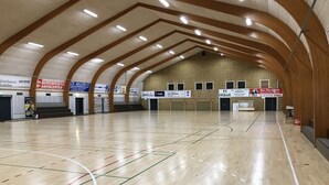 An indoor sports hall with a wooden ceiling, multiple sports courts, and various signs advertising local businesses. - Bredebro camping (Bredebro)
