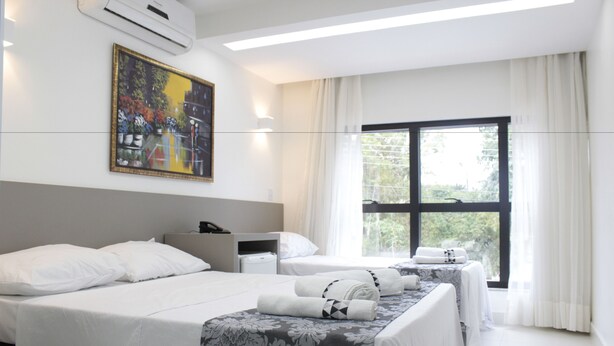 A hotel room with two beds, a painting on the wall, a window with curtains, and a bedside table with a lamp. - Hotel Hellyu's (Nucleo Bandeirante)