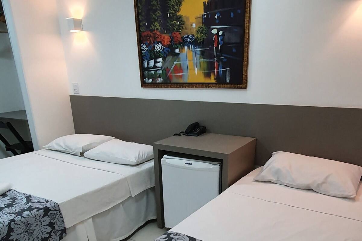 A hotel room with two beds, a small refrigerator, a painting on the wall, and an air conditioning unit.