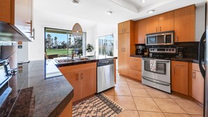 Een moderne keuken met houten kasten, roestvrijstalen apparatuur, een vaatwasser en een groot raam met uitzicht op palmbomen.