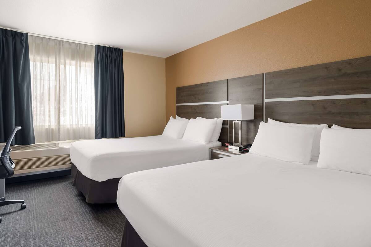 A hotel room with two beds, a desk, a chair, a lamp, and a window with curtains.