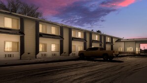 A multi-story motel with a row of rooms, a parking area, and a building with a flat roof.