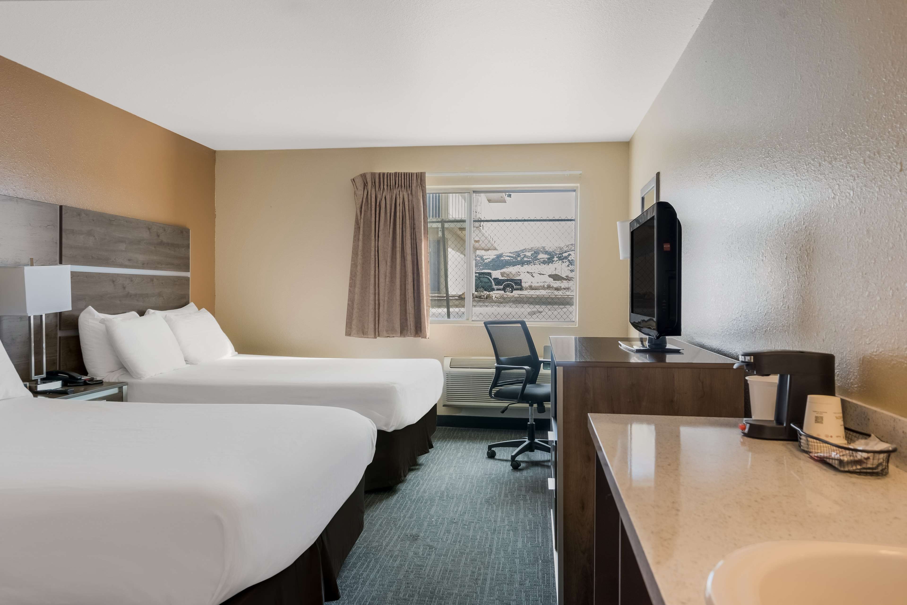 A hotel room with two beds, a desk with a computer, a TV, and a window with a view of mountains.