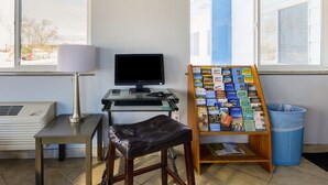 A desk with a computer, a lamp, a chair, a bulletin board with travel brochures, and a trash can.