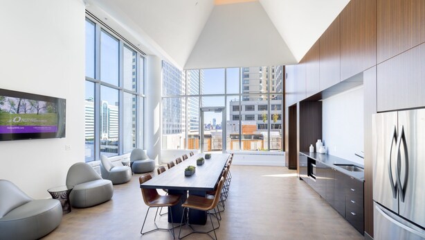 A modern kitchen with a long dining table, comfortable seating, and large windows offering city views. - Deedee's Sky Rise Apartments - Newport 2 (Jersey City)